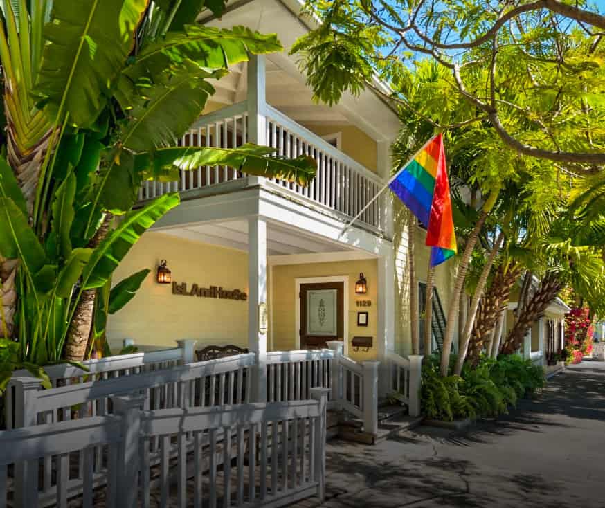 The front entrance of Island House in Key West, Florida, surrounded by lush tropical greenery. A bright rainbow flag is prominently displayed, symbolizing inclusivity and pride. The building features a light exterior, white railings, and a balcony, creating a warm and inviting atmosphere. Recognized as the world’s top gay resort, Island House is a standout LGBTQ+ friendly destination.