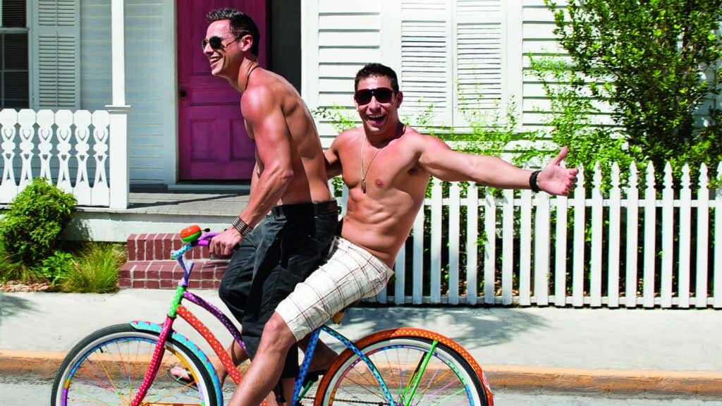 A playful and shirtless couple enjoys a sunny day in Key West, riding a colorful polka dot bike, with one sitting on the handlebars, arms outstretched, in front of a white picket fence and a pink-doored house.