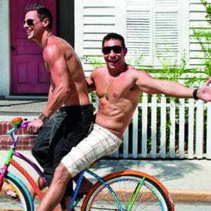 A playful and shirtless couple enjoys a sunny day in Key West, riding a colorful polka dot bike, with one sitting on the handlebars, arms outstretched, in front of a white picket fence and a pink-doored house.