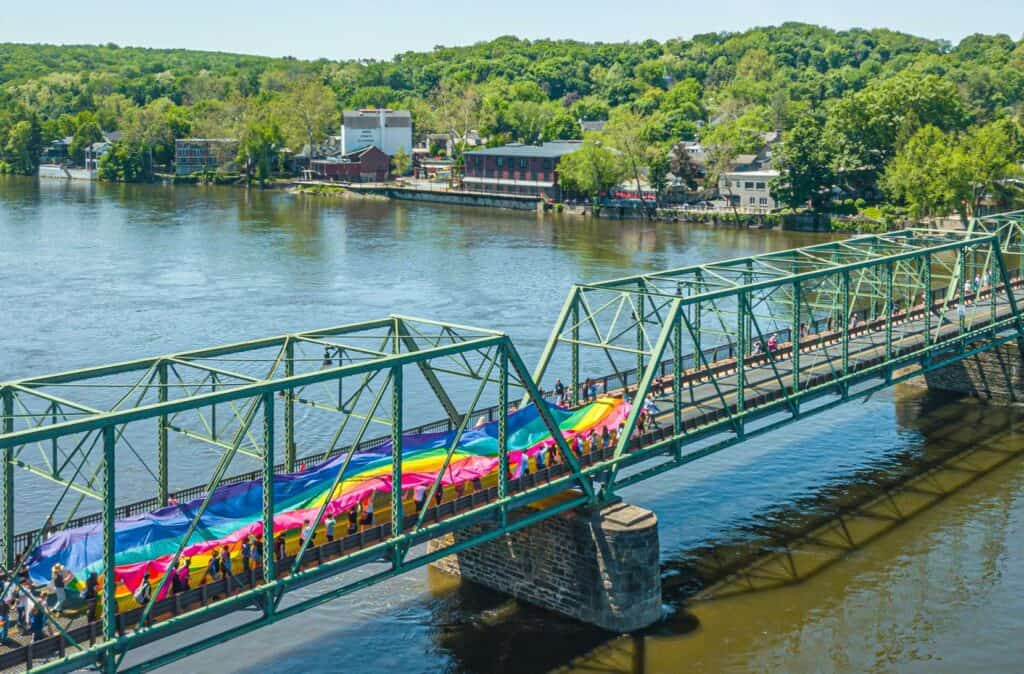 Group crossing bridge over Delaware River from Lambertville, NJ to New Hope, PA, carrying large gay pride flag - LGBTQ+ community celebrates unity and inclusion.
