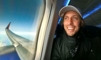 A smiling airline passenger wearing a hat and neck gaiter looks out the airplane window at the wing and blue sky during a flight.