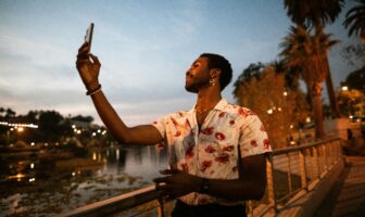 A stylish person in a floral shirt takes a selfie at sunset along a waterfront walkway lined with palm trees—an image that fits a South Beach travel guide focused on nightlife vibes.