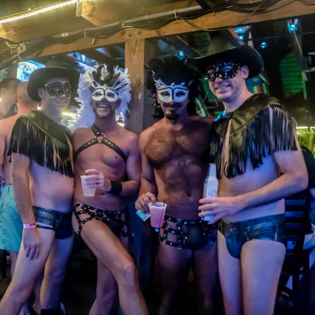 Four individuals wearing leather attire and masks socialize at a bar.