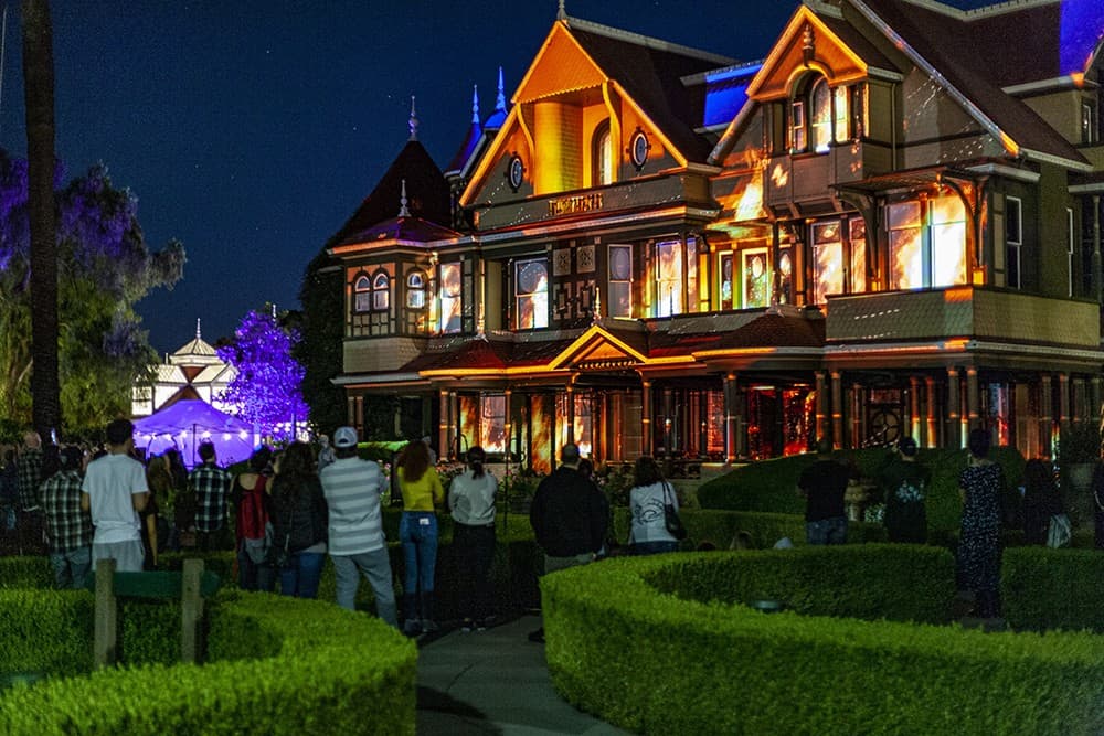 A group of individuals are standing outside the Winchester House at night, observing one of the top haunted houses in the USA.