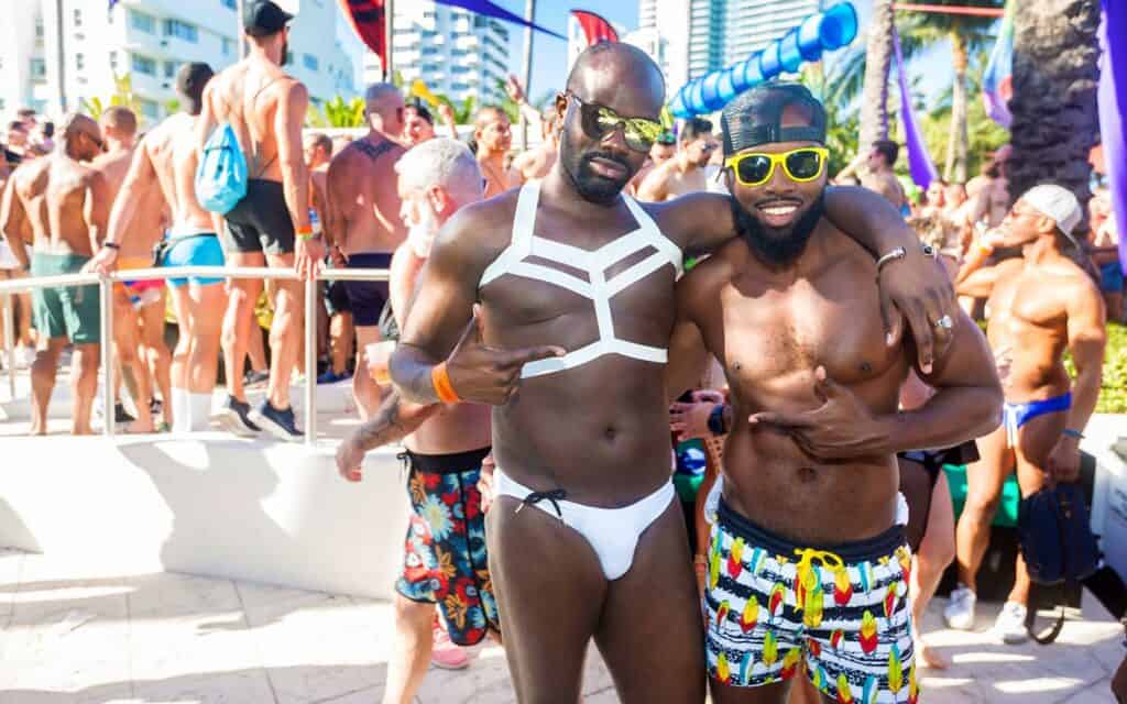 Two African American men, joyfully embracing, stand amidst the vibrant festivities of Winter Party in Miami, celebrating love and diversity.