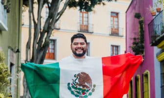 A smiling man holds the Mexican flag in a colorful street with buildings.