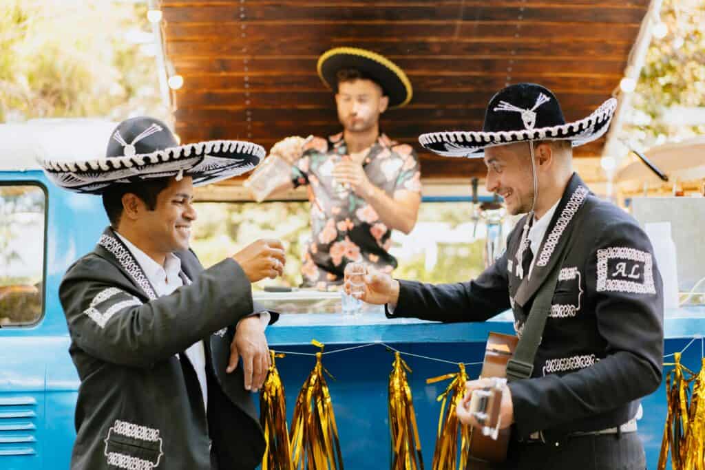 Two men in mariachi outfits toast with shot glasses during a Cinco de Mayo celebration, while a bartender in a floral shirt serves behind them.