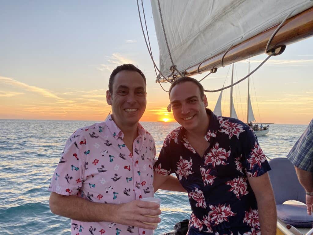 Two men from Fagabond smile aboard a sailboat during sunset in Key West; in vibrant shirts amidst calm waters with another sailboat in view.