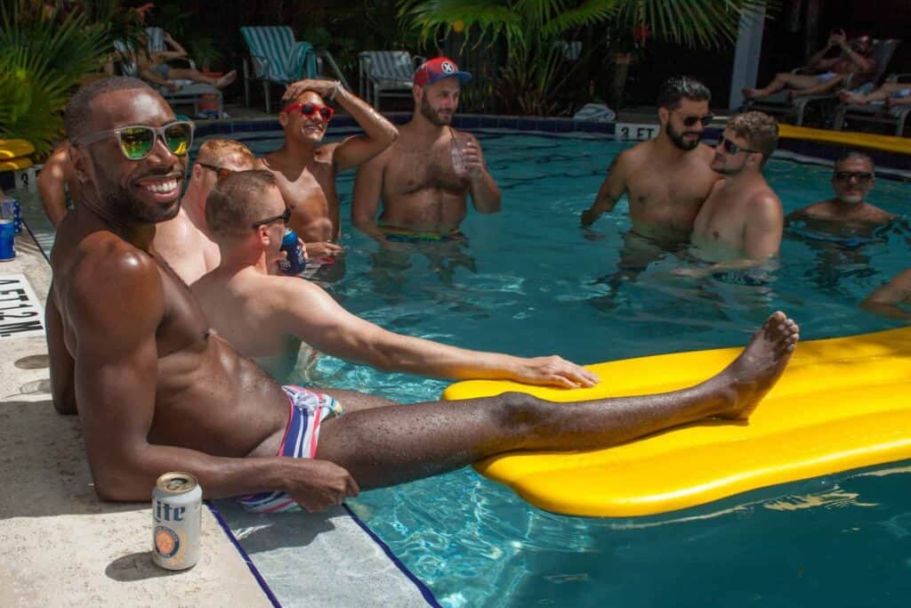 A group of men relax and socialize by the pool at Island House in Key West, Florida, surrounded by tropical greenery, with rainbow accents highlighting the welcoming and inclusive atmosphere of the world’s best gay resort.