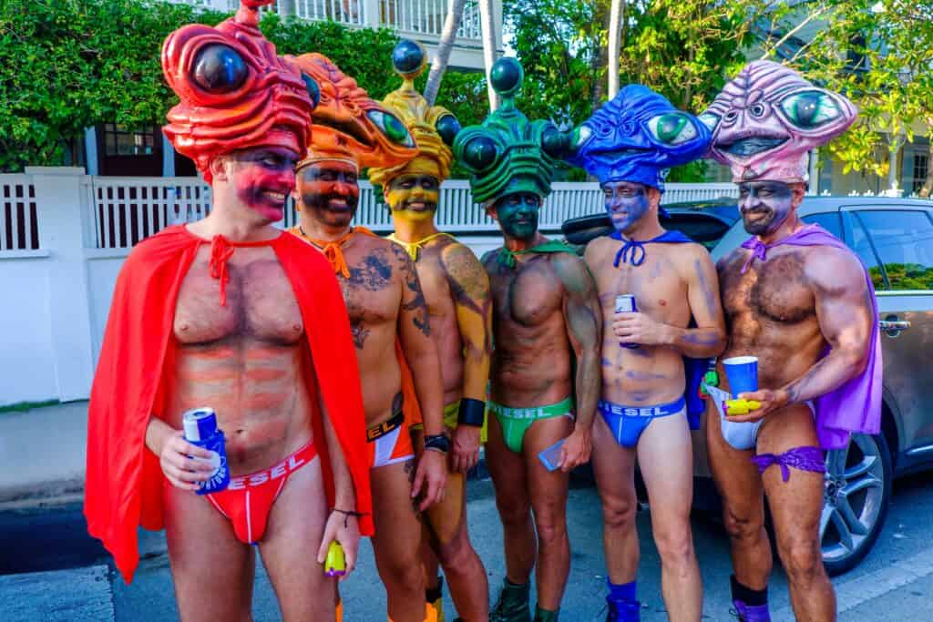 A vibrant group of six individuals dressed as rainbow-colored aliens during Fantasy Fest in Key West, Florida. Each person wears colorful, oversized alien headpieces and matching capes and underwear with "DIESEL" branding. The rainbow theme and playful outfits highlight the celebratory essence of Fantasy Fest and the inclusive vibes of Key West, making it a memorable LGBTQ+ event. Keywords: gay Key West, Fantasy Fest.