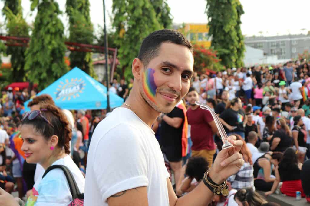 A Latino gay man smiles proudly, with a rainbow painted on his cheek, celebrating Gay Pride with joy and solidarity.