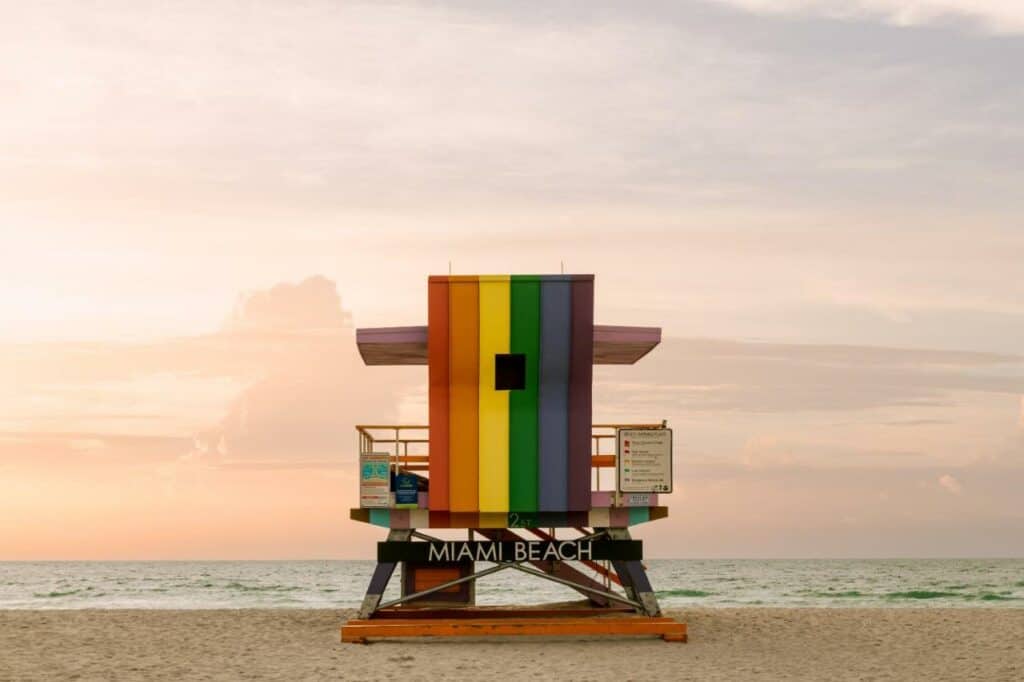 A colorful lifeguard stand painted in rainbow hues stands against the backdrop of Miami Beach, symbolizing inclusivity and diversity.