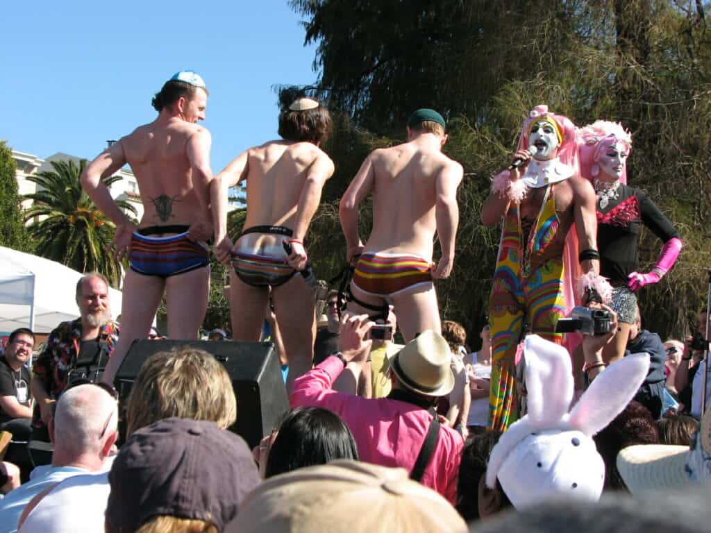 Three shirtless male contestants in underwear are observed by nuns from The Sisters of Perpetual Indulgence at the Hunky Jesus and Foxy Mary contest in San Francisco, celebrating diversity and inclusivity in the LGBTQ+ community.