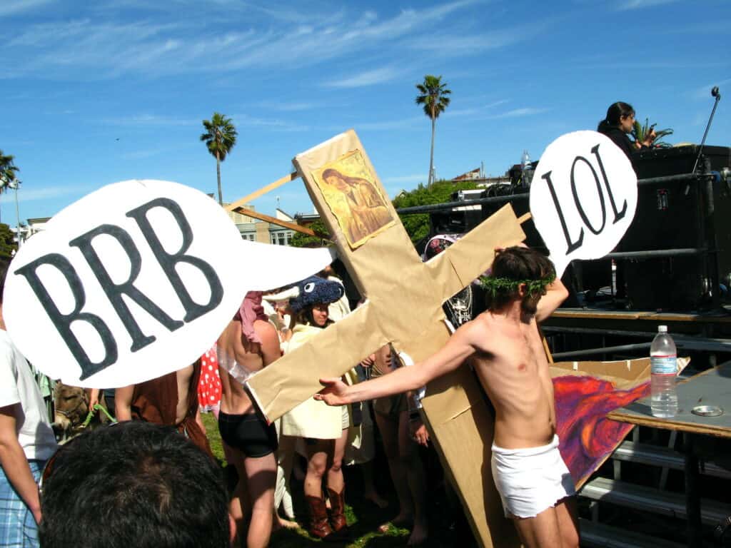 A participant dressed as Jesus on the cross with humorous "brb" and "lol" messages, surrounded by onlookers at the Hunky Jesus and Foxy Mary contest in San Francisco, celebrating diversity and inclusivity in the LGBTQ+ community.