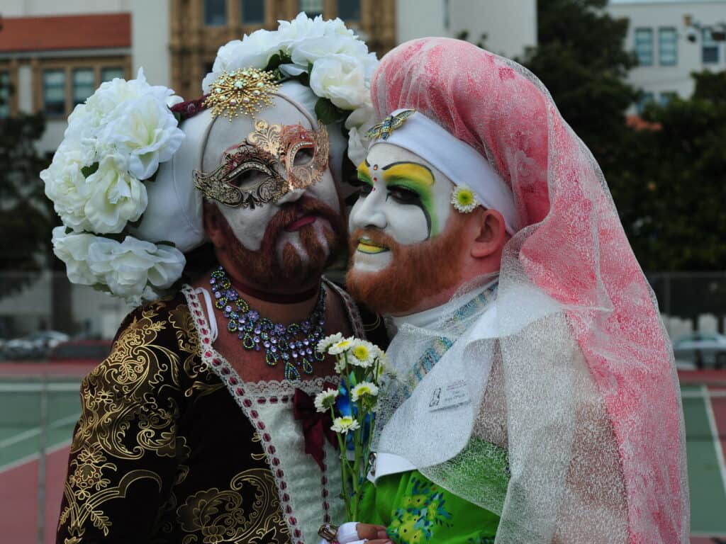 Two members of The Sisters of Perpetual Indulgence, adorned in full kubuke makeup, observe the Hunky Jesus and Foxy Mary contest in San Francisco, celebrating diversity and inclusivity in the LGBTQ+ community.