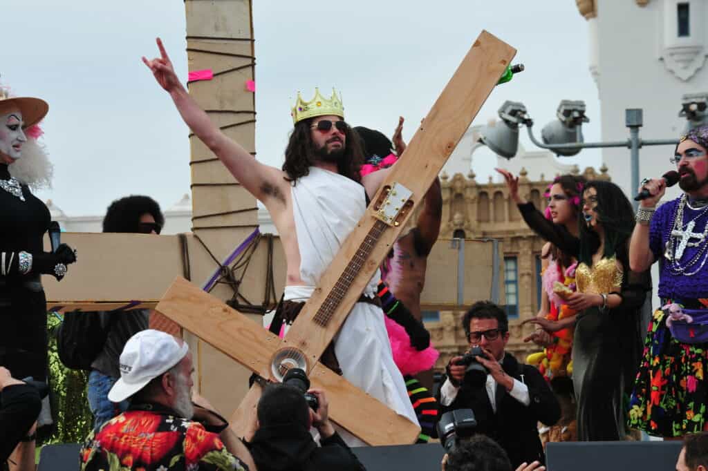 A male participant dressed as Jesus, holding a cross that resembles an electric guitar at the Hunky Jesus and Foxy Mary contest in San Francisco, celebrating diversity and inclusivity in the LGBTQ+ community.