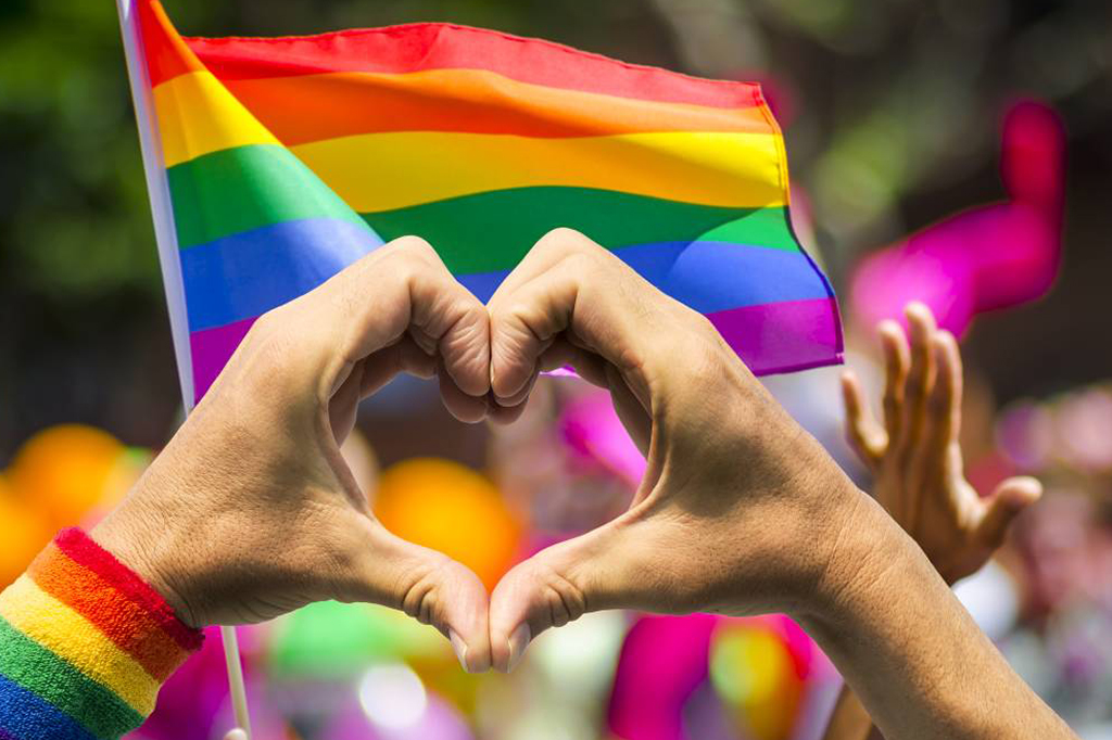 Person makes heart with hands in front of pride flag, symbolizing love and inclusivity.