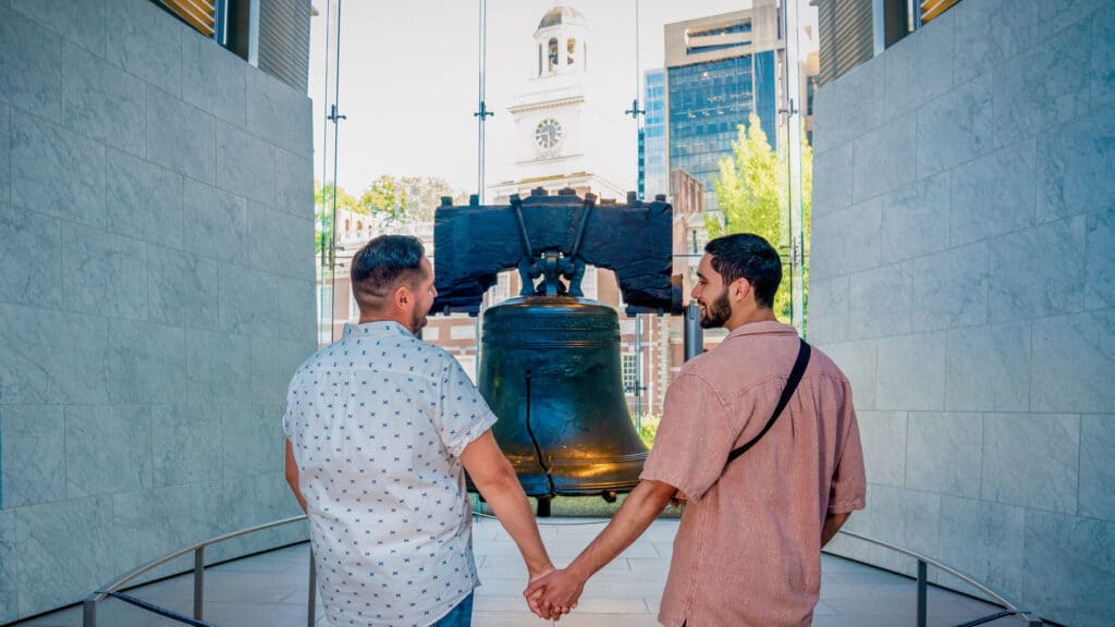 Two men holding hands in front of the Liberty Bell in Philadelphia, symbolizing love, diversity, and LGBTQ+ acceptance in the city.