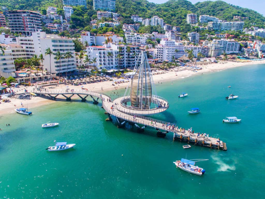 A seaside view of Los Muertos Pier in Puerto Vallarta, a popular gathering spot in the city's vibrant LGBTQ+ scene, with a large sail sculpture surrounded by boats in turquoise water.