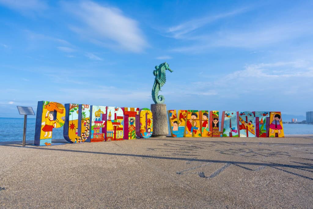 Colorful 'Puerto Vallarta' sign with a seahorse statue on a seaside promenade, celebrating the city's vibrant LGBTQ+ community and welcoming atmosphere for all visitors.