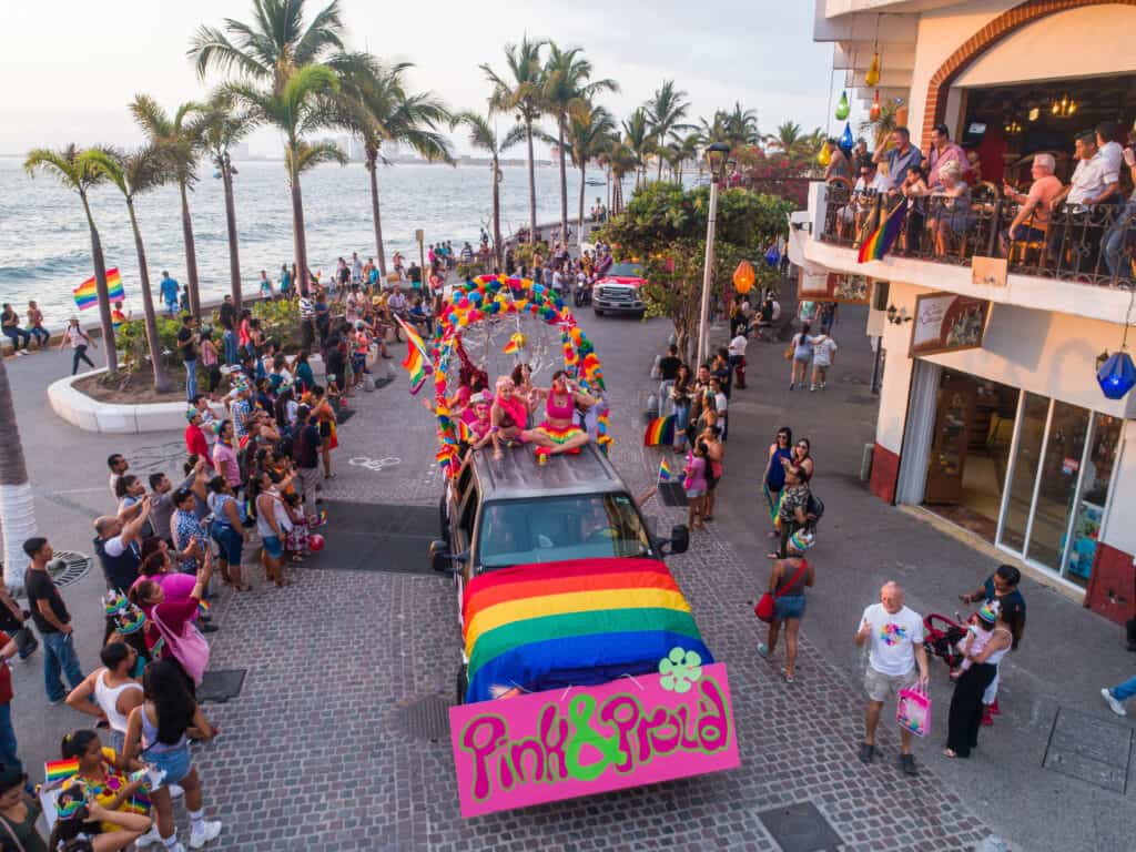 A vibrant Pride parade in Puerto Vallarta, with a decorated vehicle, rainbow flags, and a cheering crowd celebrating the city's LGBTQ+ inclusivity by the beach.