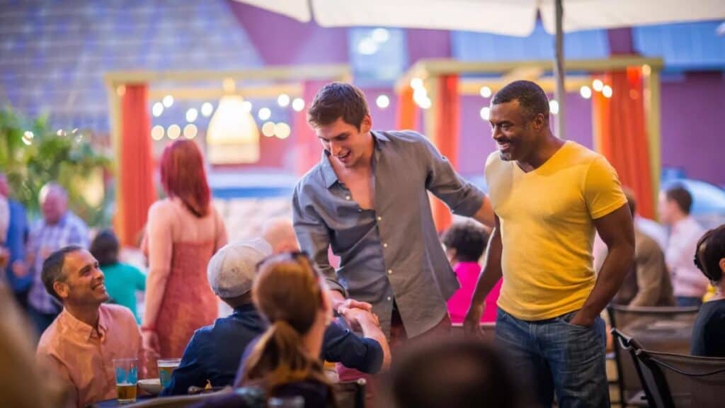 A vibrant outdoor social scene at a welcoming patio restaurant in St. Petersburg, Florida. Two men, one in a light blue shirt and the other in a yellow T-shirt, smile as they interact with others seated around a table, creating a sense of connection and community. String lights, lush greenery, and colorful accents frame the cheerful atmosphere, making it the perfect LGBTQ+ spring break destination.