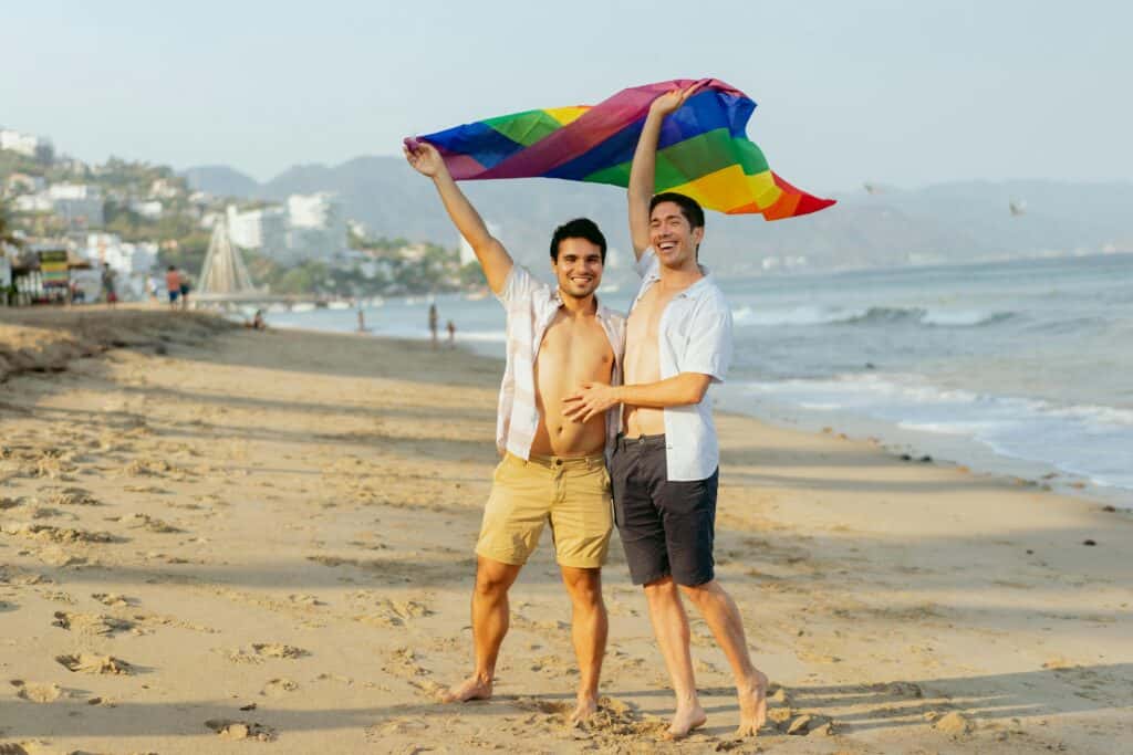 Two men celebrating on Playa de los Muertos beach in Puerto Vallarta, holding a rainbow flag and embracing the city's vibrant LGBTQ+ culture.