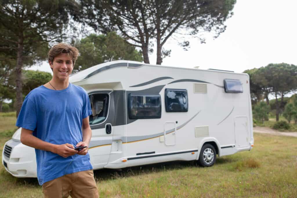 A young man stands in front of a sleek white RV with stylish black and gray patterns, set in a grassy, tree-filled location. Holding a smartphone, they smile casually, reflecting the joy of a carefree Swiftie RV road trip adventure outdoors