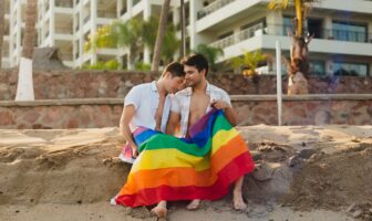 Two men sitting on a sandy beach in Puerto Vallarta, wrapped in a rainbow flag, enjoying a romantic sunset in this LGBTQ+ friendly destination.