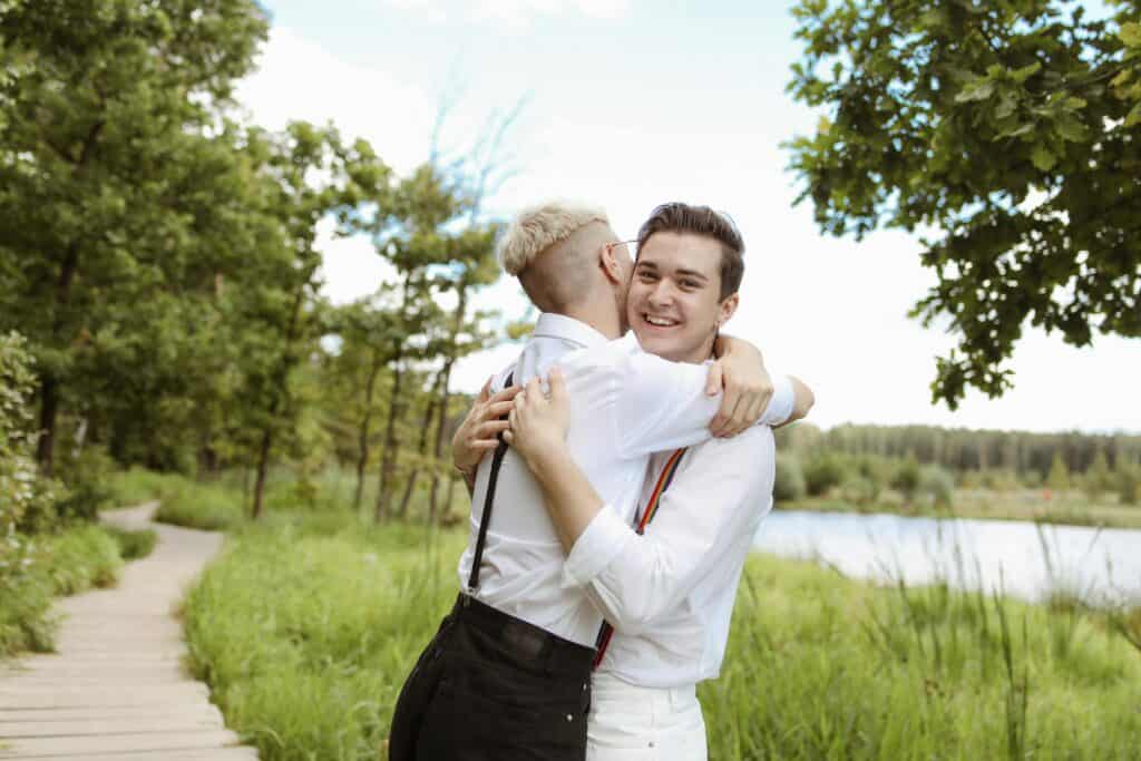 Two young men embrace happily on a scenic lakeside trail, surrounded by lush greenery, soaking in the beauty of an off-the-beaten-path LGBTQ+ Spring Break Destination—far from the crowds, yet full of adventure, romance, and unforgettable memories.