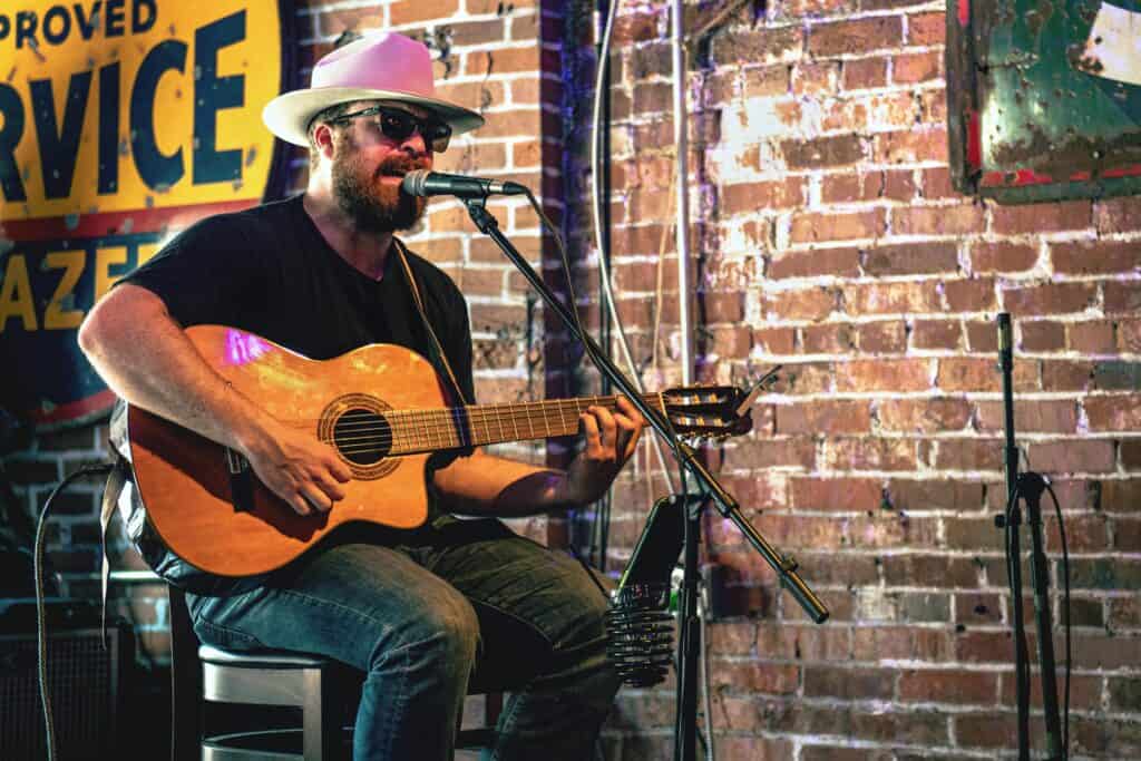 A must-see in Nashville on a Swiftie RV road trip! A musician wearing a white hat and sunglasses plays an acoustic guitar in front of a microphone. The rustic brick wall and a vintage 'SERVICE' sign in the background add to the cozy, nostalgic vibe, resembling a quintessential Nashville live music scene.