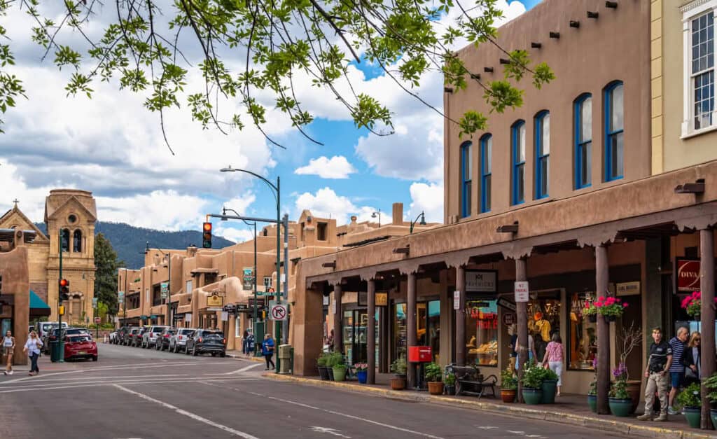 A vibrant street scene in Santa Fe, NM, with adobe-style architecture and colorful shopfronts, including signs for "101 Mavericks" and "Diva." A church with a bell tower is visible in the distance, while trees frame the sky—a lively hub known for its inclusive vibe, perfect for LGBTQ+ travelers exploring spring break destinations.