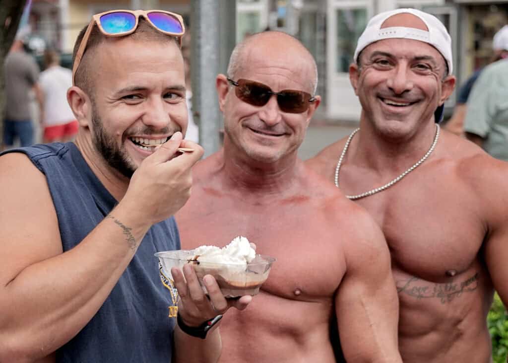 Three men smiling together, one holding a dessert bowl with whipped cream while attending a Provincetown LGBTQ+ event.