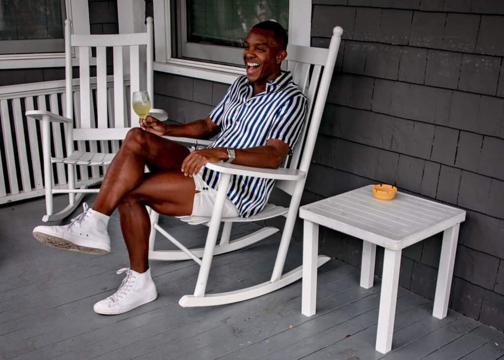 A man relaxing in a rocking chair, holding a drink and smiling, with a small table nearby.