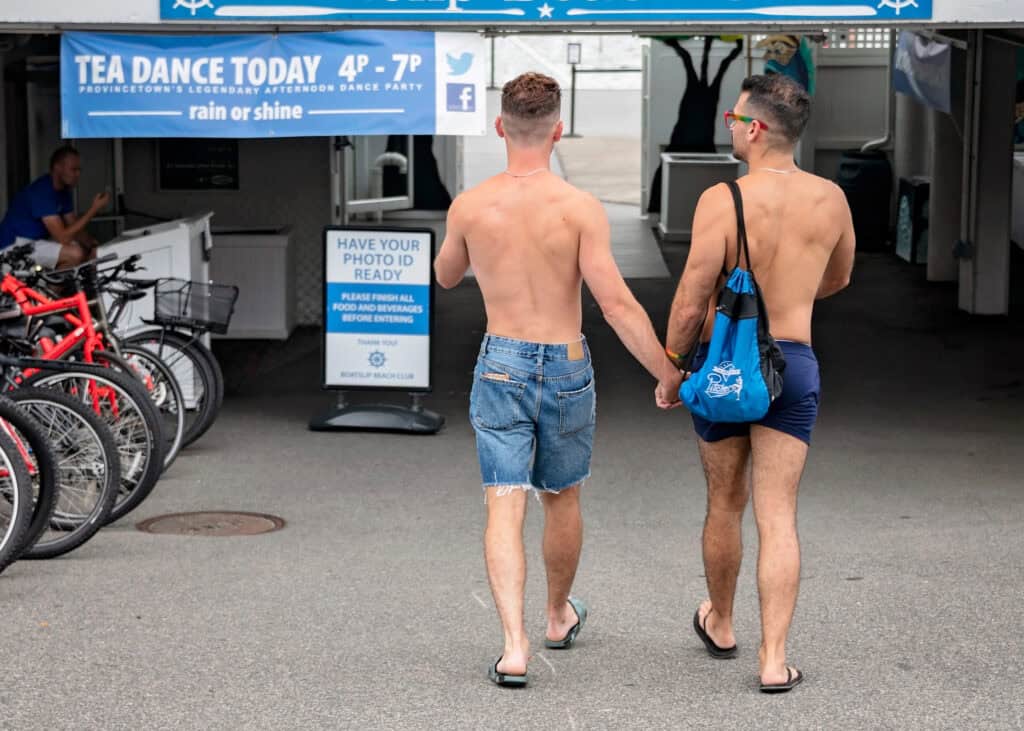 Two men walking hand-in-hand, wearing shorts and sandals, near a sign for Tea Dance at The Boatslip in Provincetown.