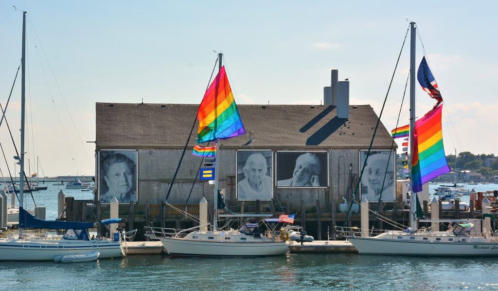 Provincetown's famous MacMillan Pier with sailboats and colorful rainbow flags, featuring large portraits on the dock's building.