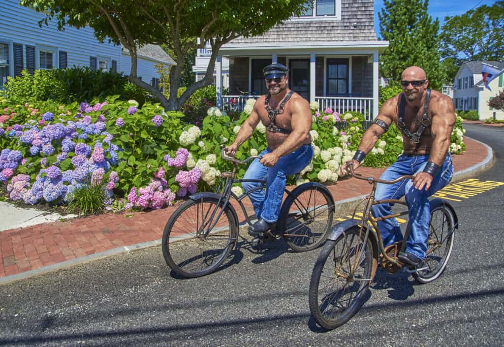 Two muscular men riding bicycles on a sunny street in PTown with colorful hydrangeas during Bear Week, a famous Provincetown LGBTQ+ event.