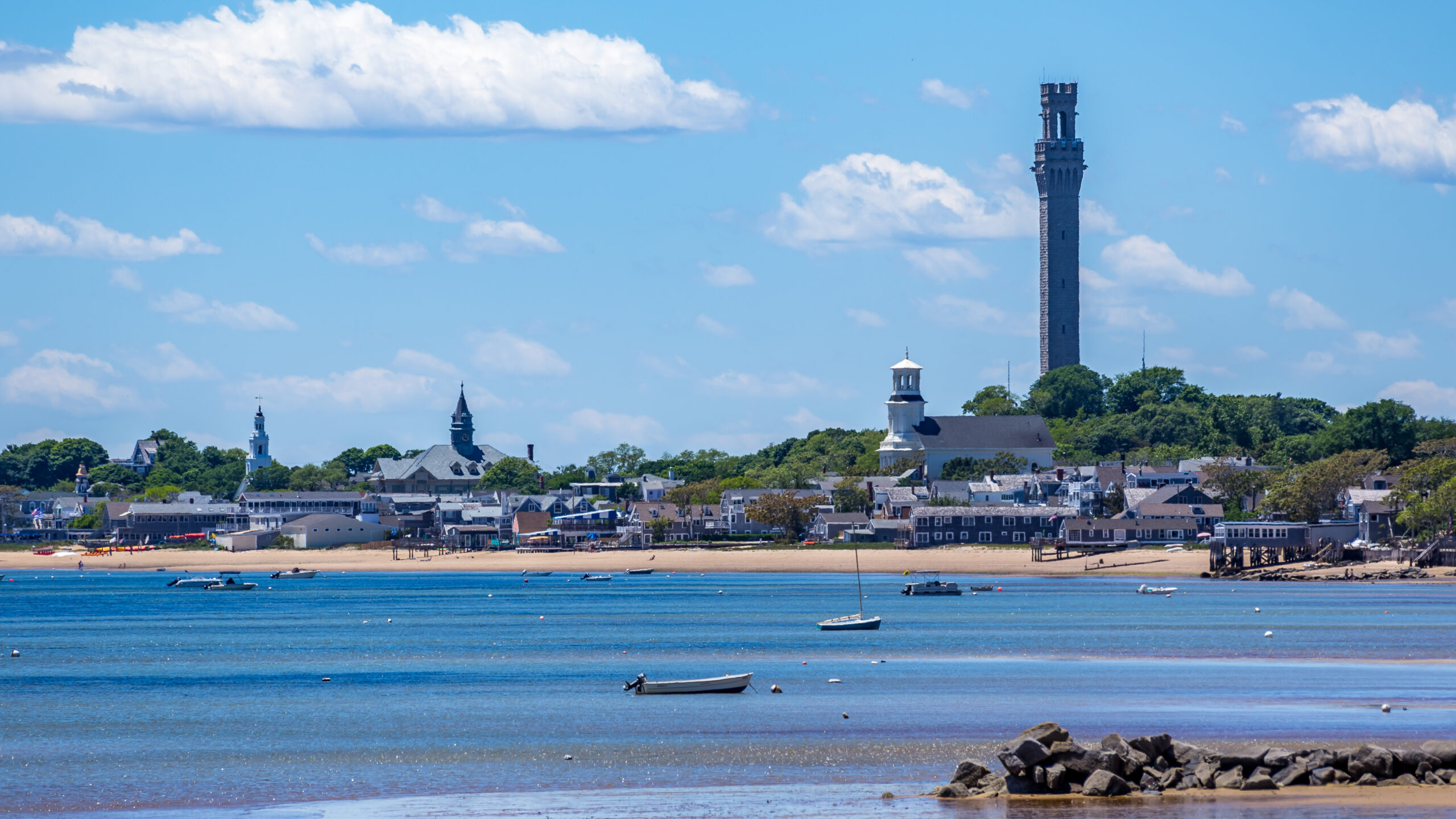 A coastal view of Provincetown with a sandy beach, boats in the water, and the Pilgrim Monument in the background.