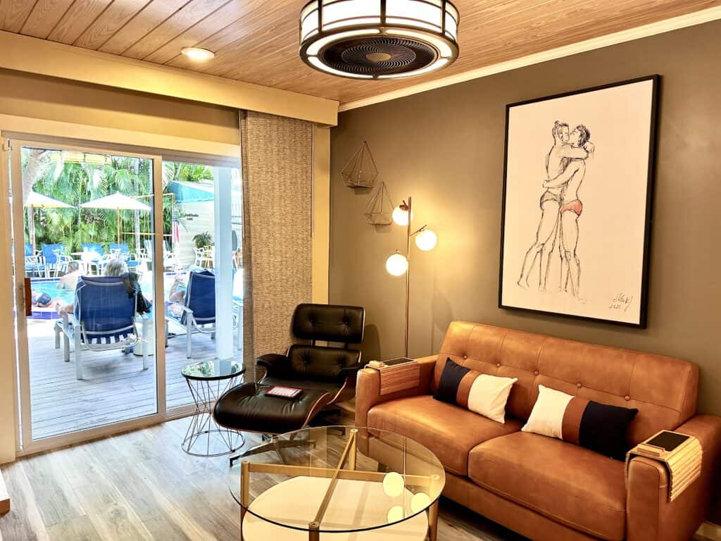Modern living room at the Island House Key West with a brown couch, glass coffee table, and artwork. Pool area visible through sliding glass door.