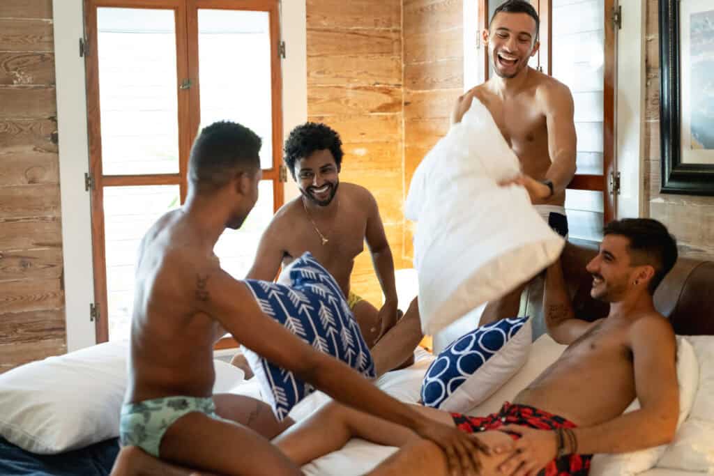 Four shirtless men playfully having a pillow fight on a bed in a cozy room at the Island House Key West.