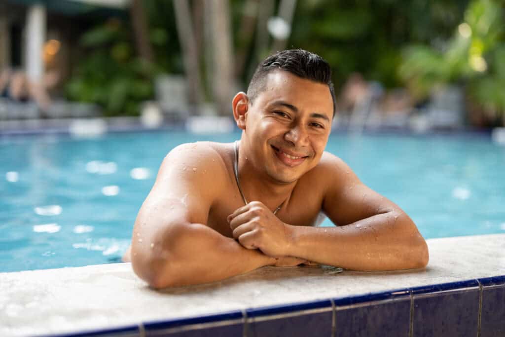 A smiling young man resting on the edge of the pool at Island House Key West, with water droplets on his arms.
