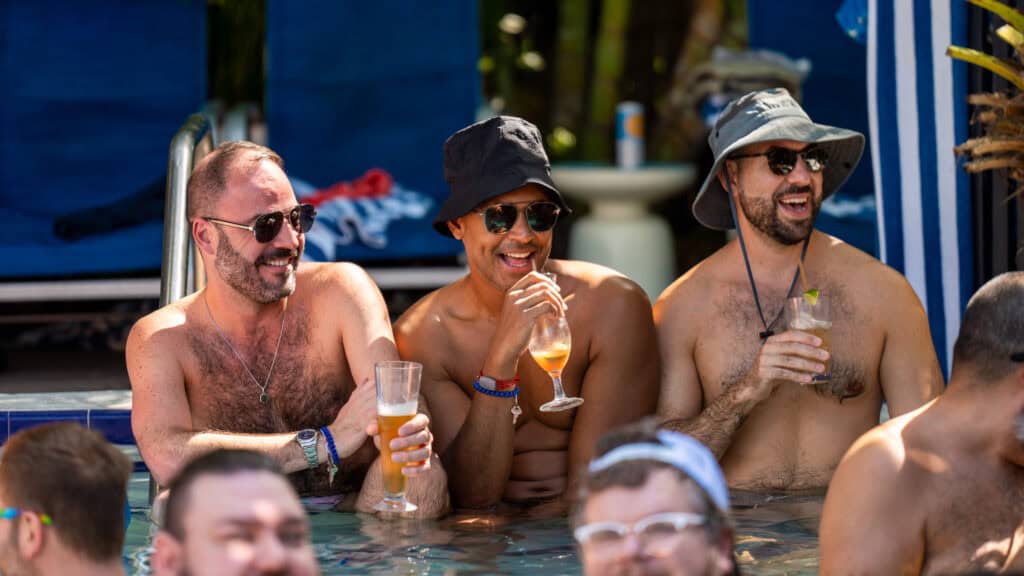 Three men sitting in a pool, enjoying drinks and smiling. They wear sunglasses and hats.