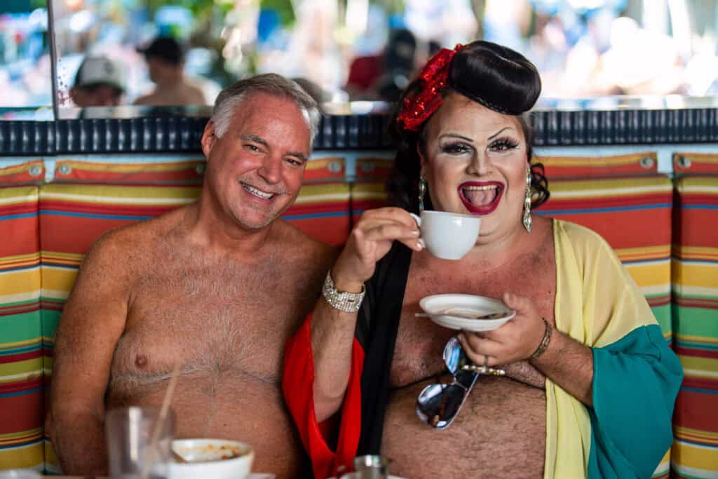 Two smiling people, one in a colorful outfit and makeup, enjoying drinks at a café.