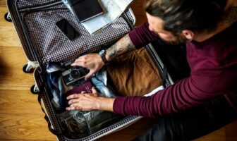 A man packing a suitcase with clothes, a camera, and a passport — all part of his gay travel packing essentials — sitting on a wooden floor.