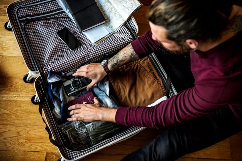A man packing a suitcase with clothes, a camera, and a passport — all part of his gay travel packing essentials — sitting on a wooden floor.
