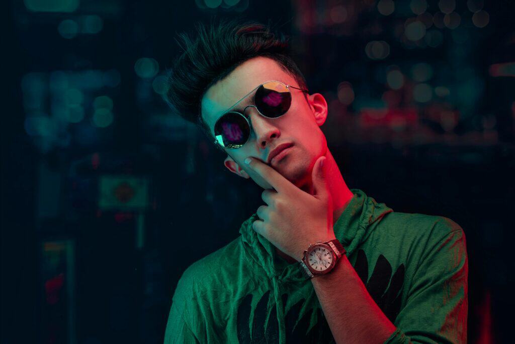 A young gay man in a green hoodie with sunglasses — a standout piece among his gay travel packing essentials — striking a thoughtful pose against a dark, colorful background.
