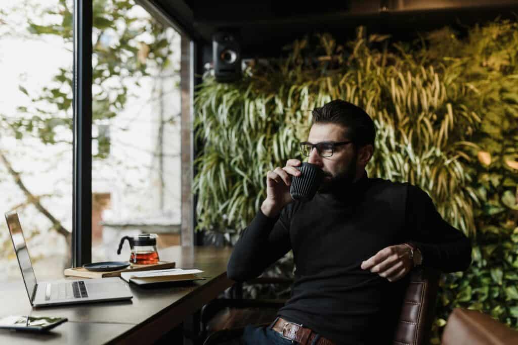 A man in a black mock turtleneck sipping from a black cup, sitting at a wooden table with a laptop.