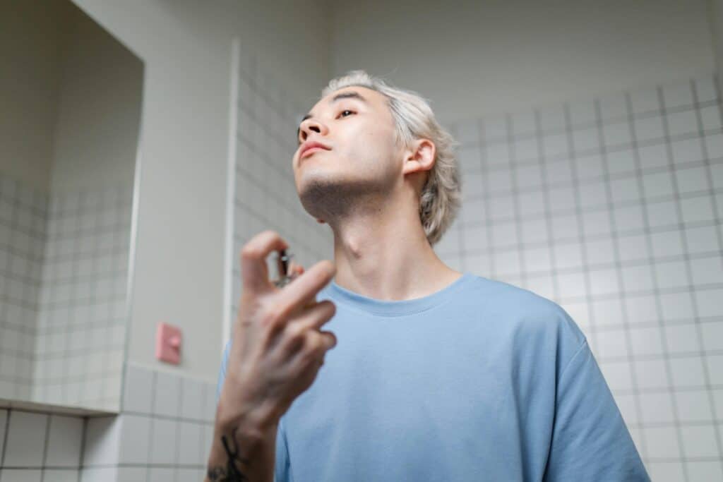 A gay man with short platinum hair sprays cologne while looking upward in a bathroom.