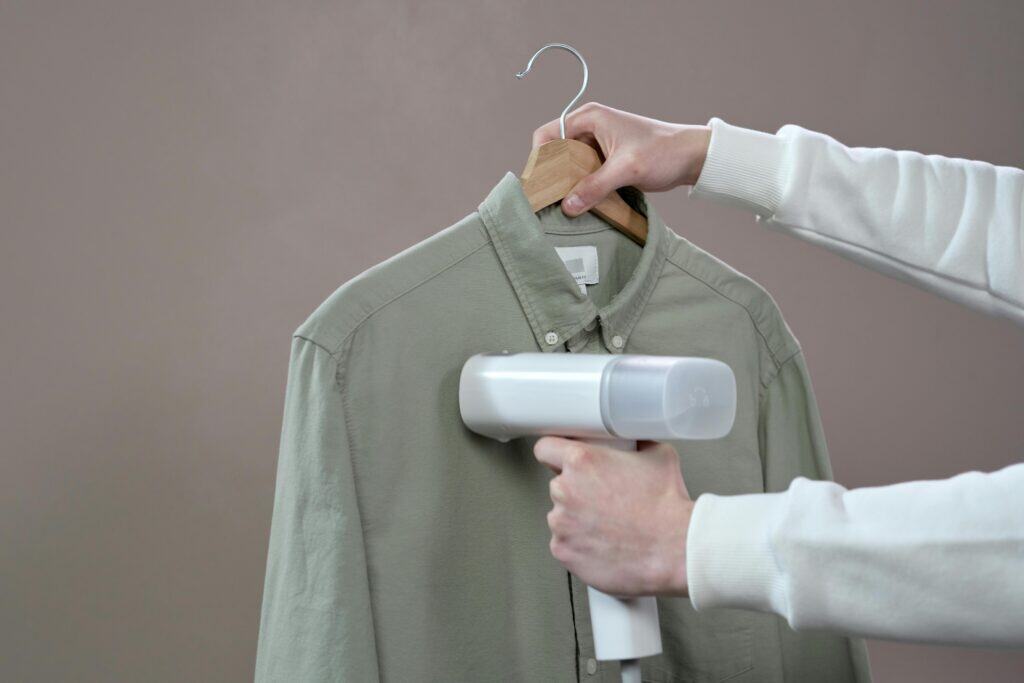 A person holding a handheld garment steamer on a green shirt hung on a wooden hanger.