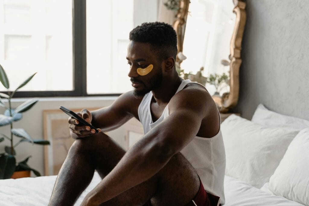 A man sitting on a bed wearing eye patches — one of his go-to gay travel packing essentials — while looking at his phone.